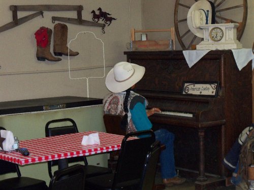A cowboy mannequin plays an old piano in a rustic room with western decor and a checkered tablecloth.