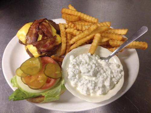 A plate with a cheeseburger topped with bacon, lettuce, tomato, pickles, crinkle-cut fries, and a side of cottage cheese.