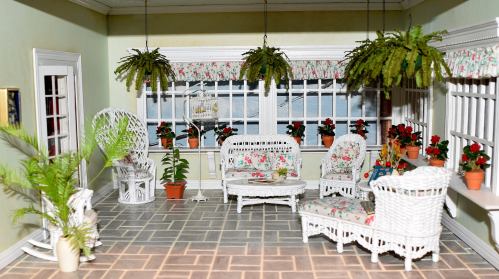 A cozy dollhouse living room with white wicker furniture, floral cushions, potted plants, and large windows.