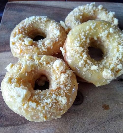 Four glazed donuts topped with crumbled cookie pieces, arranged on a wooden surface.
