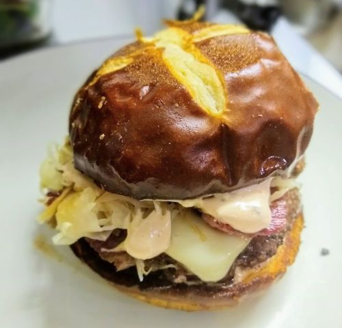 A close-up of a pretzel bun burger with layers of meat, cheese, sauerkraut, and creamy sauce on a white plate.