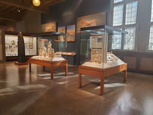 Museum interior featuring display cases with ancient artifacts and sculptures, illuminated by natural light.
