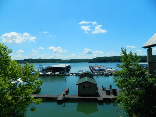 A serene lake view with boats docked, surrounded by green trees and a clear blue sky with fluffy clouds.