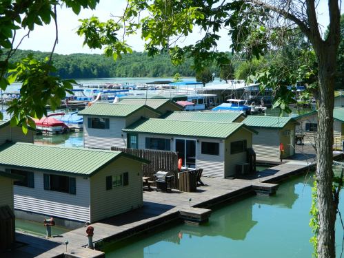 Aerial view of houseboats on a calm lake, surrounded by greenery and a marina with boats in the background.