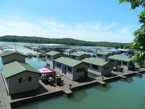 A scenic view of houseboats on a calm lake, surrounded by green hills and a marina in the background.