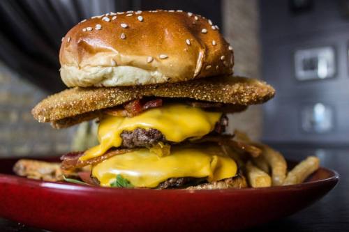 A towering burger with two beef patties, melted cheese, bacon, and a crispy layer, served with fries on a red plate.