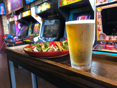 A frosty beer glass next to a plate of tacos, with vintage arcade games in the background.