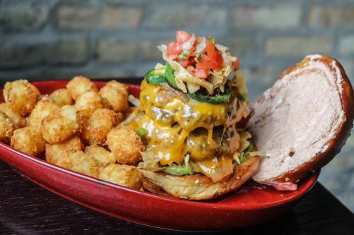 A towering cheeseburger topped with jalapeños and salsa, served with crispy tater tots on a red plate.