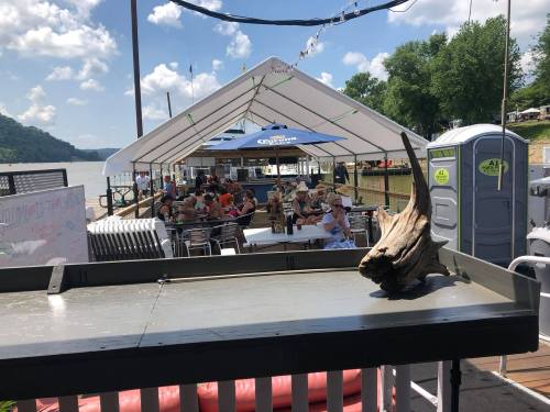 A riverside venue with a large tent, tables filled with people, and a decorative driftwood piece in the foreground.