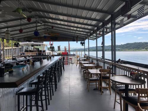 A waterfront restaurant with a bar and tables, featuring a covered patio and scenic views of the water and hills.
