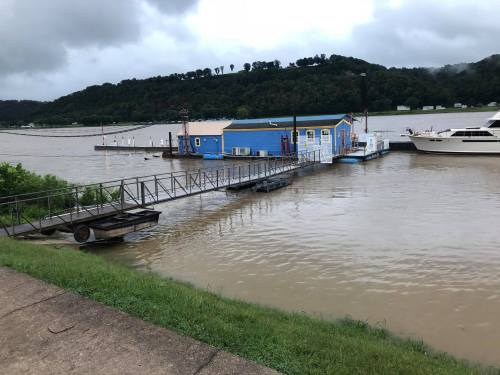 A blue houseboat docked at a flooded riverbank, surrounded by water and green hills under a cloudy sky.