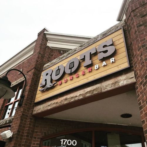 Sign for "Roots Burger Bar" on a brick building, featuring bold letters and a wooden background.