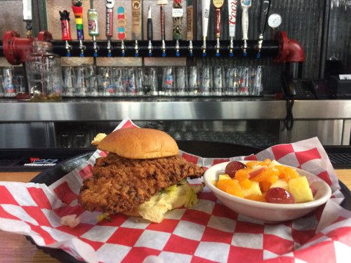A crispy fried chicken sandwich on a bun with lettuce, served with a side of fruit salad in a bowl.