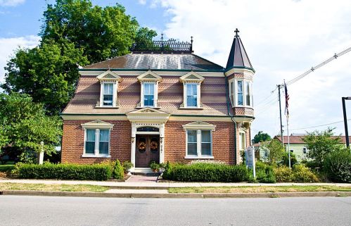 A charming two-story brick house with a pointed turret, surrounded by greenery and a clear blue sky.