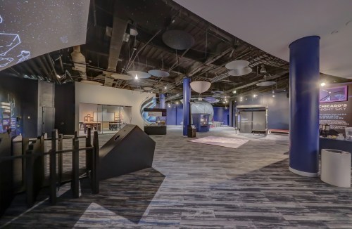 A spacious, modern exhibit area with dark walls, blue columns, and various interactive displays about the night sky.