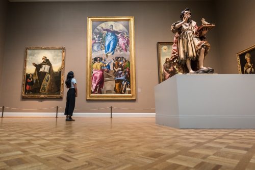 A person stands in an art gallery, admiring large paintings and a statue on display.