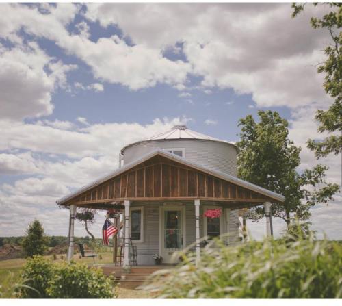 A charming round house with a porch, surrounded by greenery and under a cloudy sky, featuring an American flag.