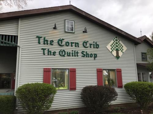 Exterior of a building with green signage reading "The Corn Crib The Quilt Shop," surrounded by shrubs and a cloudy sky.