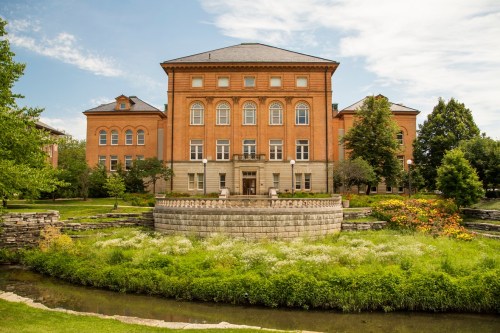 Historic brick building surrounded by greenery and a small pond, under a clear blue sky.