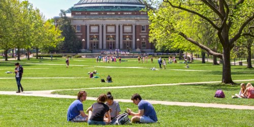 Students gather on a grassy area, enjoying a sunny day with a historic building in the background.