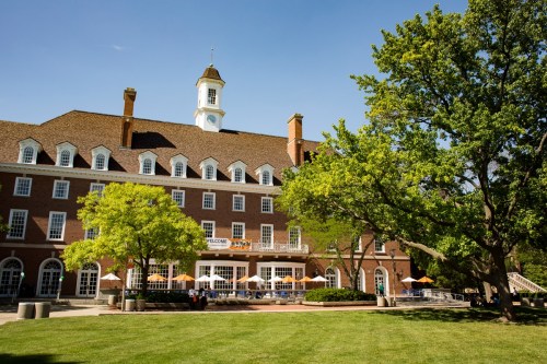 A brick building with a clock tower, surrounded by green trees and a lawn, under a clear blue sky.