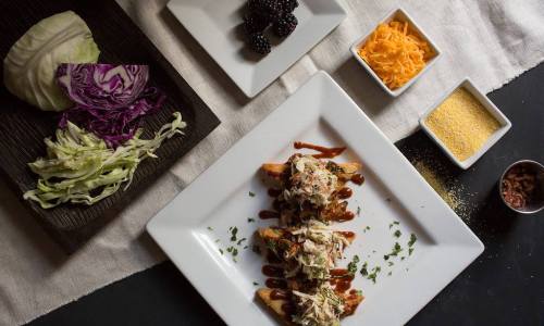 A plate of nachos topped with shredded chicken, drizzled with sauce, alongside cabbage, cheese, blackberries, and cornmeal.