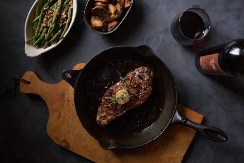 A cast iron skillet with a cooked steak, surrounded by roasted potatoes, green beans, and a glass of red wine.