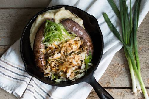 A cast iron skillet with two sausages on mashed potatoes, topped with sautéed cabbage and green onions.
