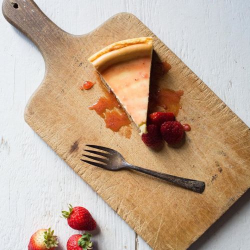 A slice of cheesecake on a wooden board, garnished with strawberries and raspberries, with a fork beside it.