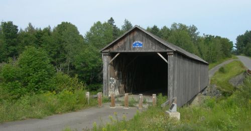 A rustic wooden covered bridge surrounded by greenery, with a gravel path leading up to it.