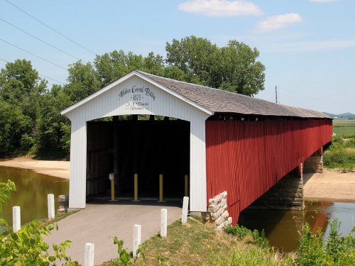 A red covered bridge spans a calm river, surrounded by greenery and blue skies. The year 1875 is visible on the bridge.