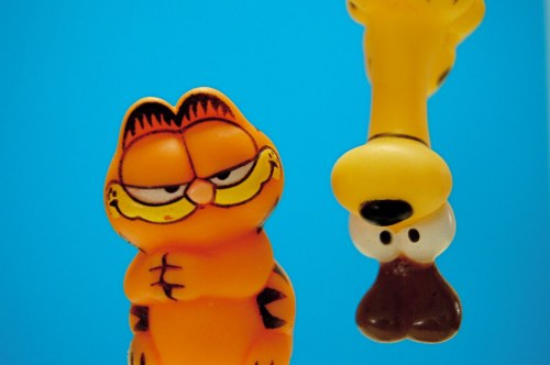Two cartoon figures: a smirking orange cat and a yellow dog, set against a bright blue background.