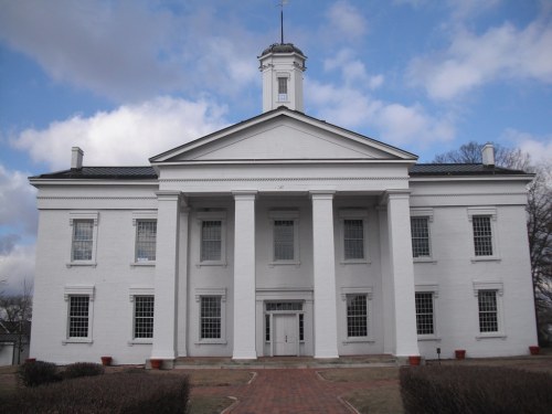 A large white building with columns and a clock tower, set against a cloudy sky. Pathway leads to the entrance.