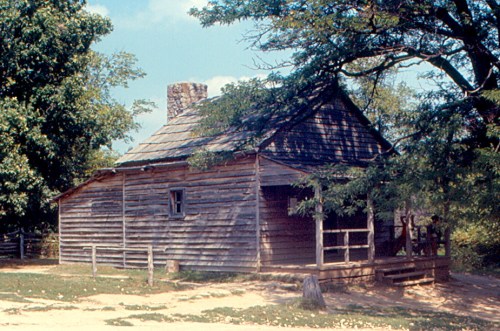 A rustic wooden cabin surrounded by trees, featuring a porch and a stone chimney under a clear blue sky.