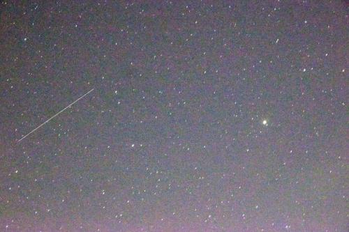 A starry night sky filled with numerous stars, featuring a bright object and a faint streak, possibly a meteor.