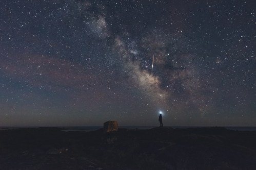 A silhouette of a person standing on a rocky landscape, gazing at a starry sky with the Milky Way and a shooting star.