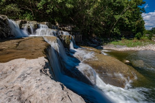 A serene waterfall cascading over rocky terrain, surrounded by lush greenery and a calm river.