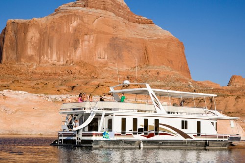 A houseboat on calm water near a large red rock formation, with people enjoying the sun on the deck.