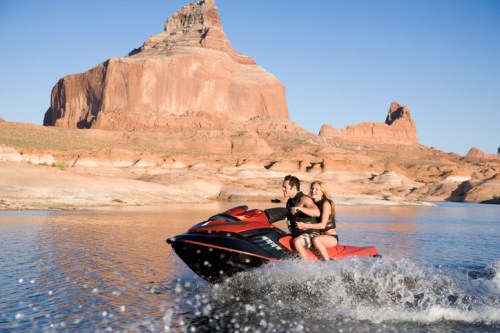 A man and woman ride a jet ski on a lake, with red rock formations in the background under a clear blue sky.