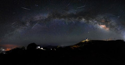 A panoramic view of the night sky filled with stars and meteors, with a person standing on a hilltop.
