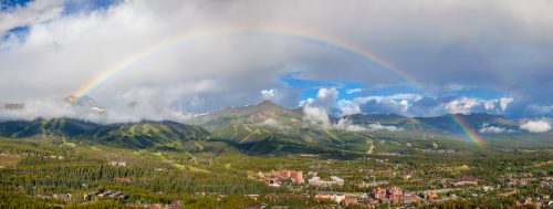 A panoramic view of a lush valley with a rainbow arching over mountains and a small town below, under a partly cloudy sky.