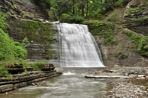 A serene waterfall cascades over rocky cliffs, surrounded by lush greenery and a calm river below.