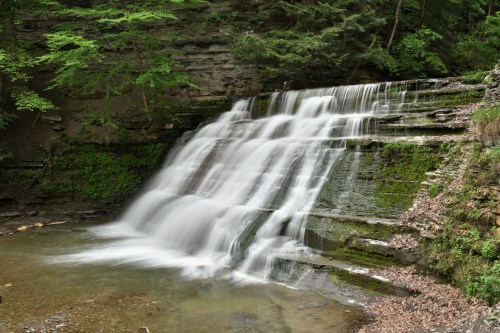 A serene waterfall cascades over moss-covered rocks into a calm pool, surrounded by lush green trees.