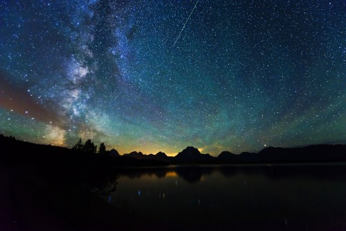 A stunning night sky filled with stars and the Milky Way, reflecting on a calm lake with mountains in the background.