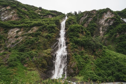 A tall waterfall cascades down a lush green mountainside, surrounded by dense vegetation and rocky terrain.