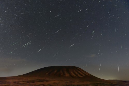 A night sky filled with shooting stars over a dark, volcanic landscape under a starry backdrop.
