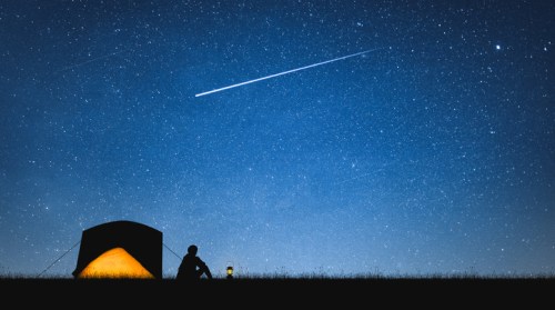 A silhouette of a person sitting by a tent under a starry sky, with a shooting star streaking across.