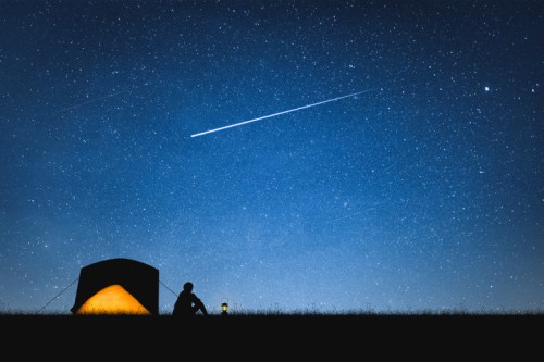 A person sits beside a tent under a starry sky, with a bright meteor streaking across the night.