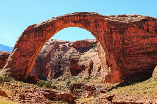 A large natural rock arch stands majestically against a clear blue sky, surrounded by rugged red rock formations.