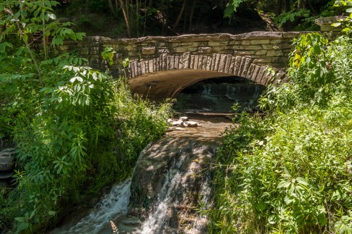 A stone bridge arches over a small waterfall, surrounded by lush greenery and trees.